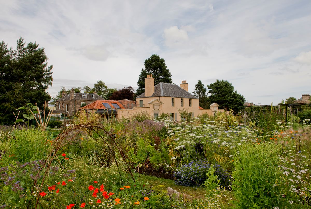 Garden with lots of greenery and red and white flowers, and a cottage in the background.