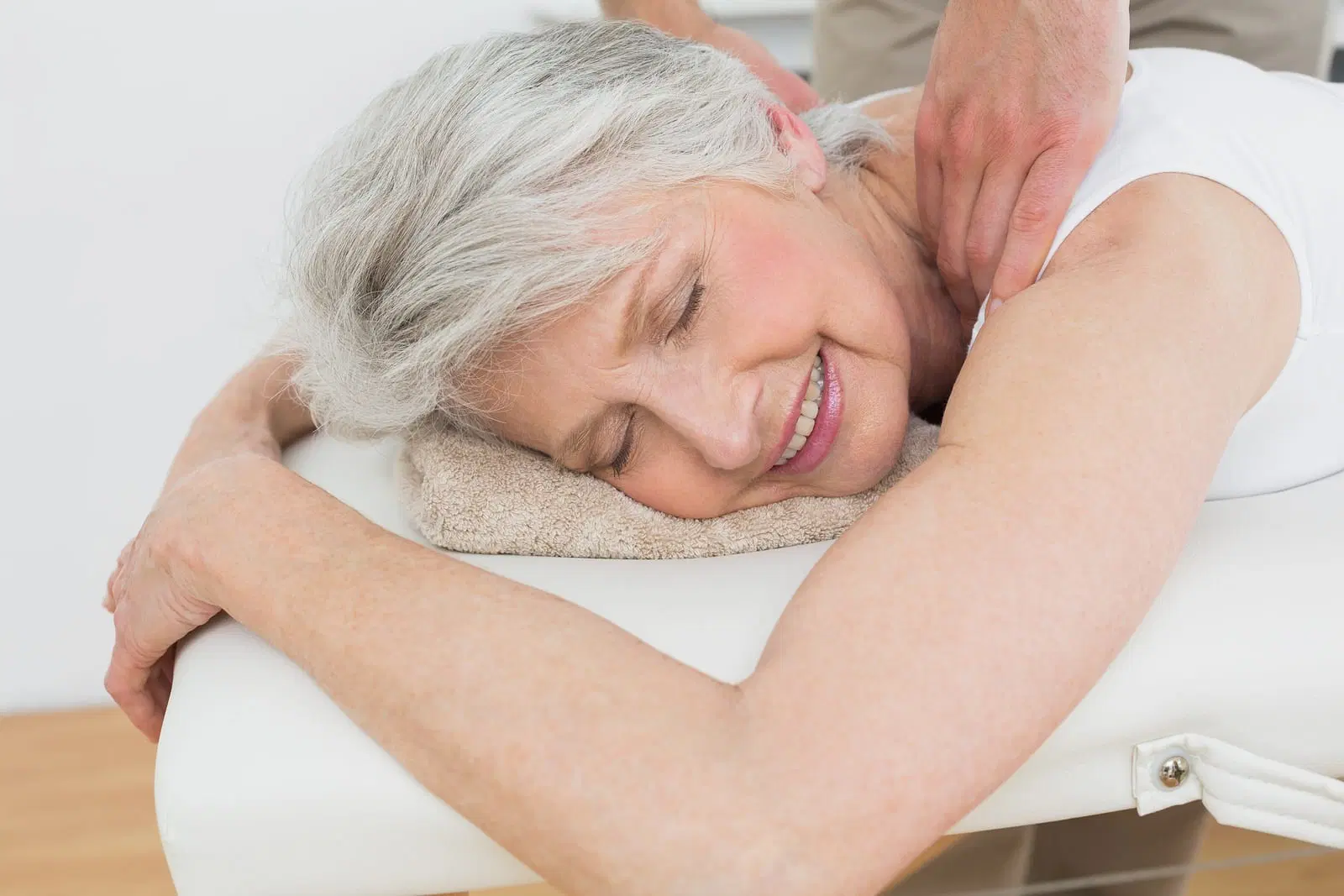 older white woman with grey hair lying down and receiving a shoulder massage and smiling.