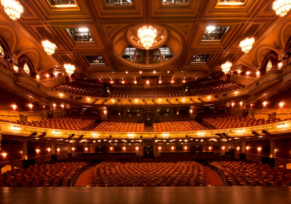View of a large theatre, from the middle of the stage. All the seats are empty but golden lights are on all around the room.